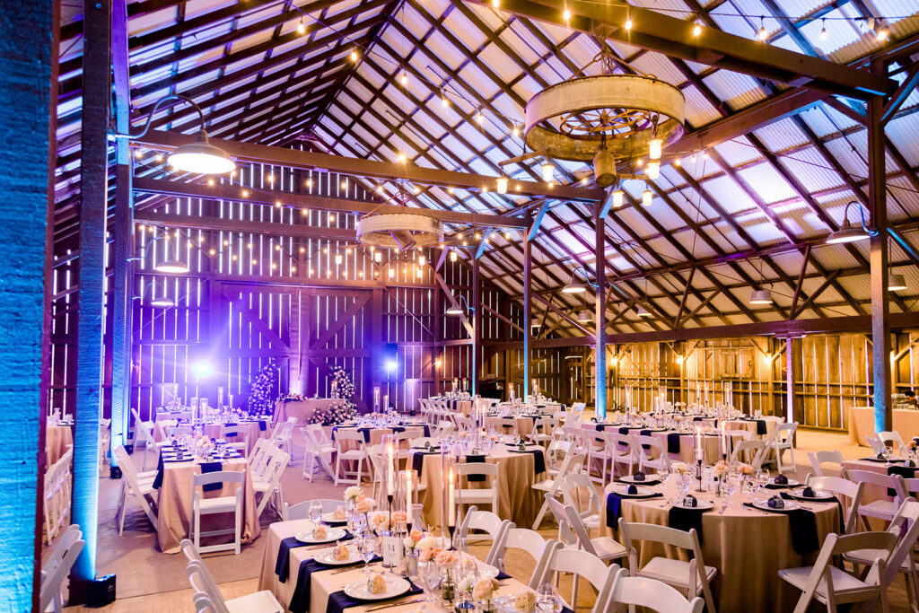 A picturesque white barn at the Slo Reception Site illuminated with beautiful blue uplighting. The barn is surrounded by a serene landscape, creating a charming and atmospheric setting for events