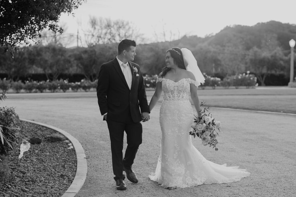 A timeless black and white image capturing a bride and groom strolling together during the golden hour at the picturesque white barn in Slo. The couple is bathed in warm, soft sunlight, creating a romantic and enchanting scene against the rustic backdrop.