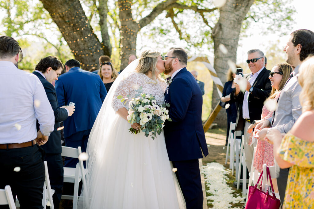 A heartwarming moment captured in an image as the bride and groom share a passionate kiss in the middle of the aisle, sealing their union and joyously exiting as husband and wife. The love and happiness between the newlyweds radiate in this beautiful post-ceremony scene