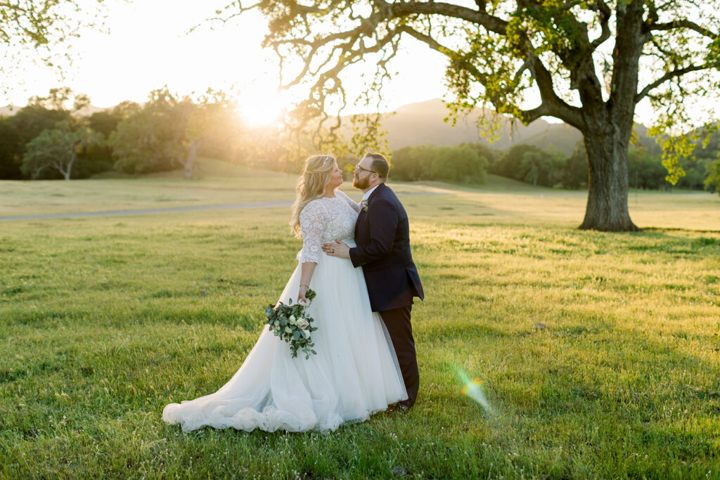 A breathtaking image of a bride and groom in a green field at Spanish Oaks Ranch, bathed in the golden light of the sunset. The couple, silhouetted against the warm hues of the evening sky, shares an intimate moment in the picturesque setting, capturing the romance of their special day